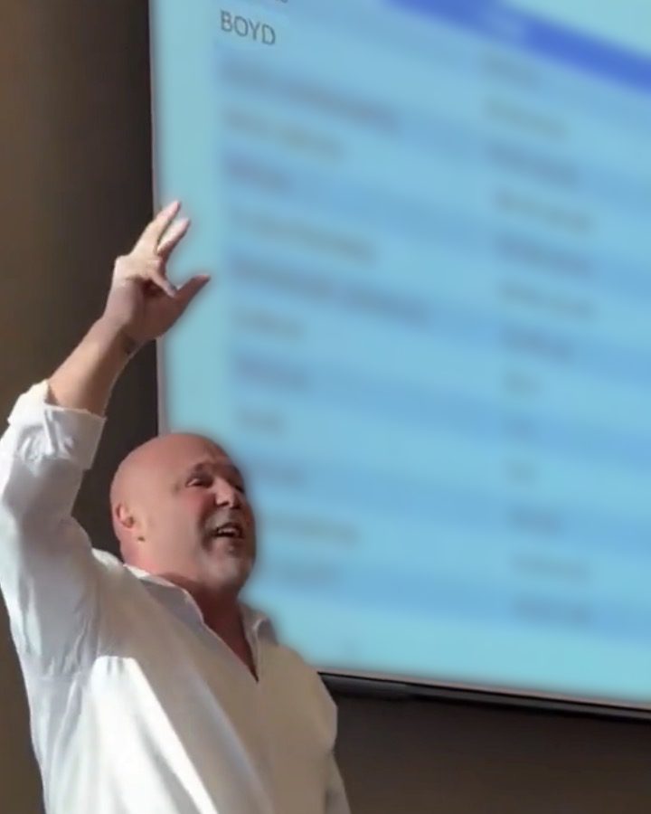 Boyd Myers on stage, one arm raised mid-talk, with the day’s speaker agenda on the screen behind him listing BOYD first